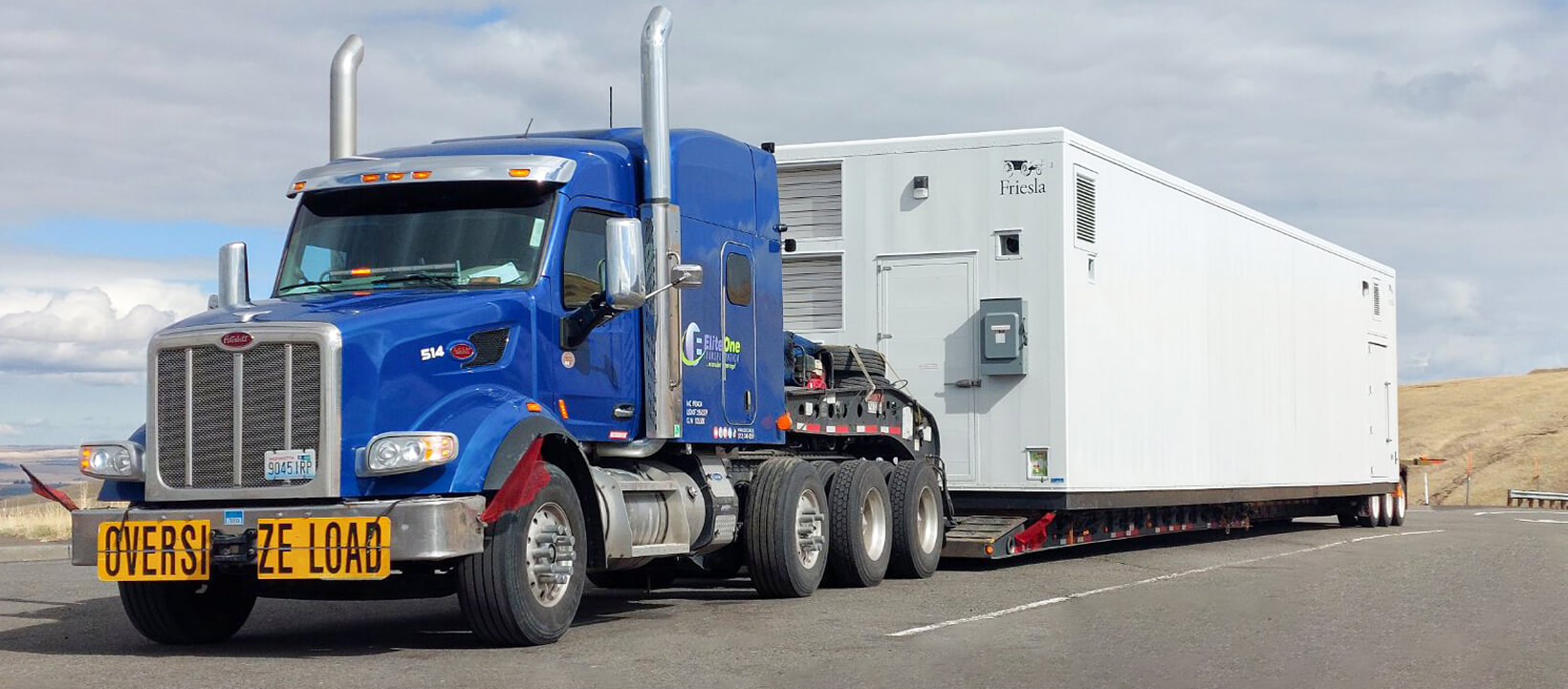 Blue Elitone Transportation truck hauling a large modular unit on a flatbed trailer marked as "Oversize Load".