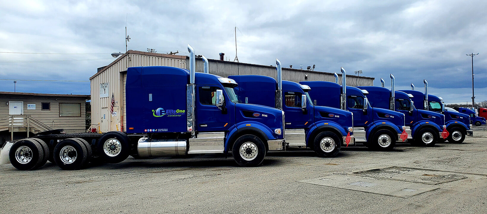 Lineup of blue Elitone Transportation semi-trucks parked in front of company facility under cloudy skies.