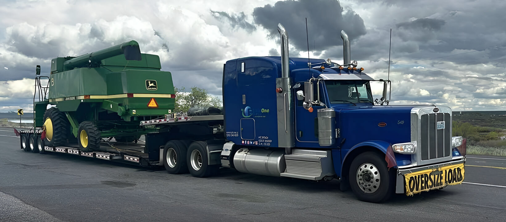 Blue Elitone Transportation truck transporting a large green John Deere harvester on a flatbed trailer marked “Oversize Load.”